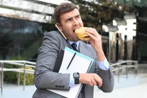 überarbeiteter Mann der unterwegs telefoniert und Fast Food isst