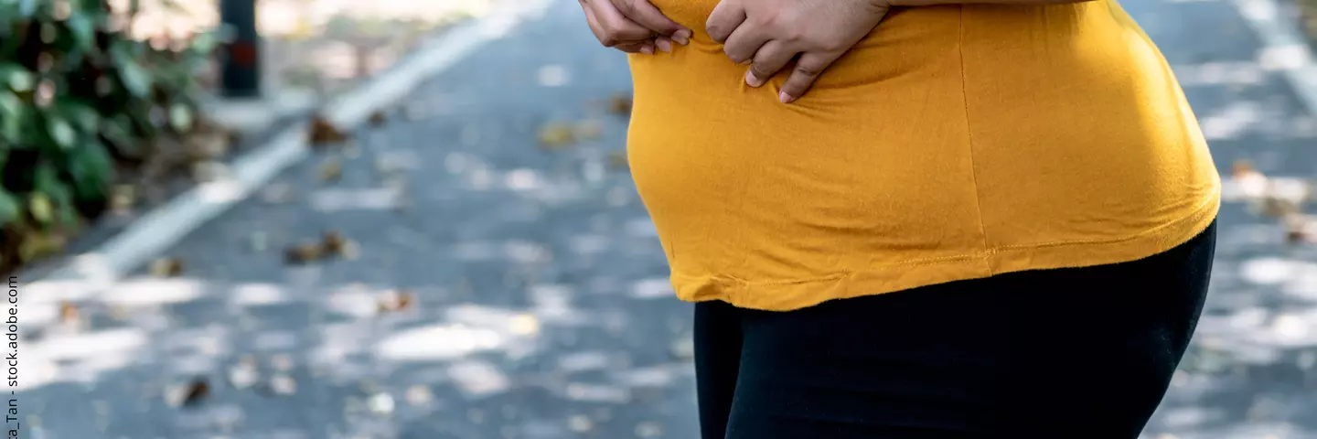 Frau mit fülliger Figur in gelbem T-shirt steht im Park und fasst sich mit beiden Händen an den Bauch